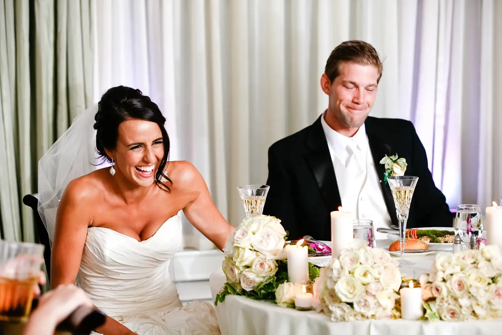 A bride laughing during a wedding speech as the groom smiles beside her, seen from where the speaker is standing